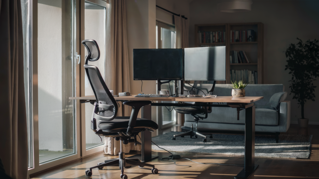 A modern home office with the best desk placement, featuring a dual monitor setup on a desk by a large window, with sunlight, a gray sofa, and a tall plant.