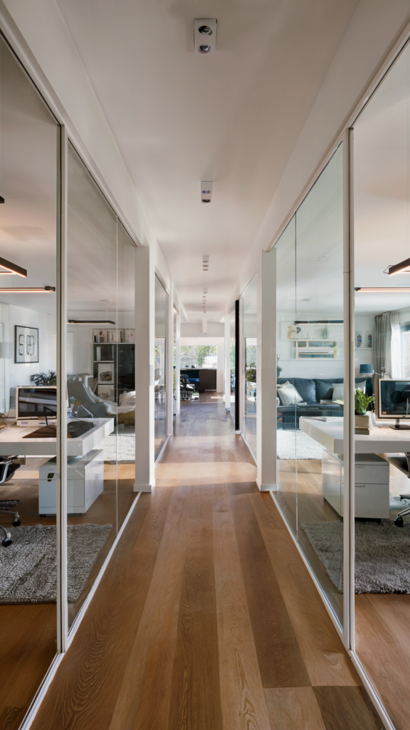 A modern office space with glass partitions separating workspaces, polished wooden floors, neutral color palette, and a blend of work and living areas in an open-concept layout.