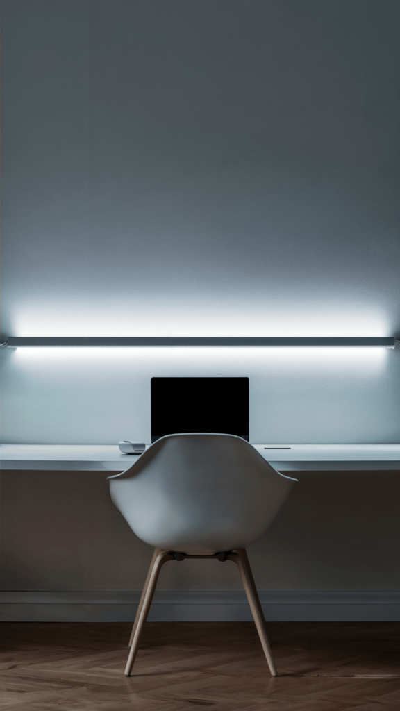 A minimalist desk with a linear white light fixture casting a soft glow over a modern white chair and laptop, set against a light blue wall.