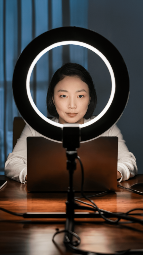 A woman illuminated by a circular ring light, highlighting her features as she works on a laptop in a dimly lit professional setting.