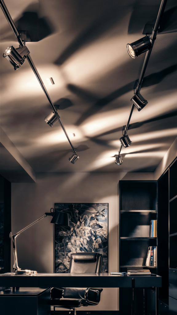 A modern office with dynamic track lights casting shadows over a dark desk, featuring a black leather chair and an abstract painting on the wall.